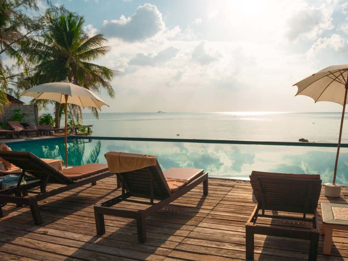 Lounge chairs and umbrellas are arranged on a wooden deck by an infinity pool, overlooking a calm sea and distant horizon under a partly cloudy sky.