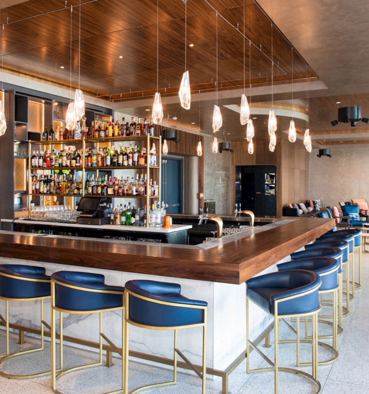 A modern bar interior with a central island, blue bar stools, hanging lights, and a well-stocked liquor shelf in a spacious setting.