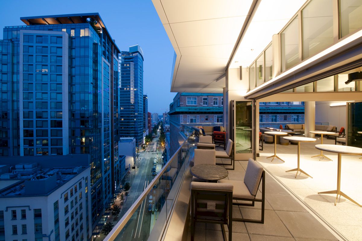 A modern balcony with seating overlooks a cityscape at twilight, featuring tall buildings and illuminated streets.