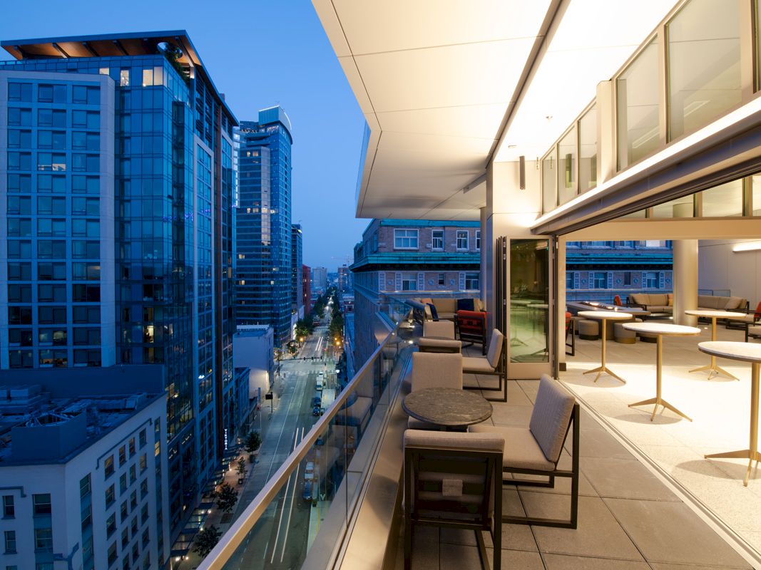A modern balcony with seating overlooks a cityscape at twilight, featuring tall buildings and illuminated streets.