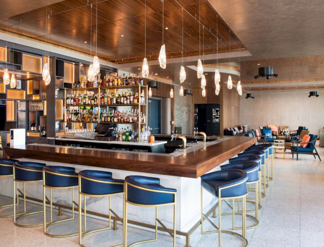 A modern bar with a wooden counter, stylish blue and gold stools, hanging lights, and a well-stocked drink selection.