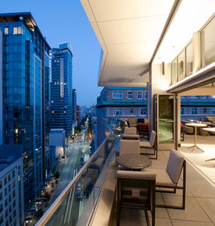 A high-rise building rooftop with outdoor seating overlooks a city skyline at dusk, showing tall buildings and lit streets below.
