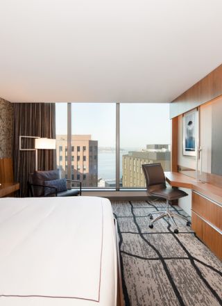 A modern hotel room features a large bed, desk with a chair, and a flat-screen TV. The window offers a cityscape and waterfront view.