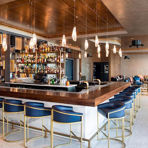 A modern bar with blue chairs, wooden accents, pendant lights, and a well-stocked shelf. Lounge seating and neutral decor complete the space.