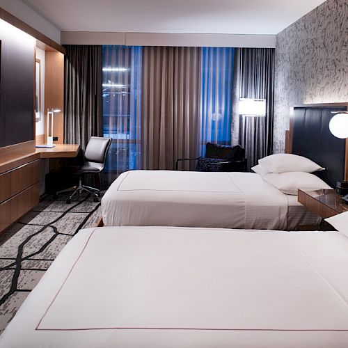 A modern hotel room with two beds, a desk, a flat-screen TV, and stylish decor featuring patterned walls and curtains.