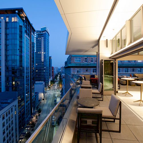 A modern rooftop balcony overlooking a city street at dusk, featuring seating areas and ambient lighting.