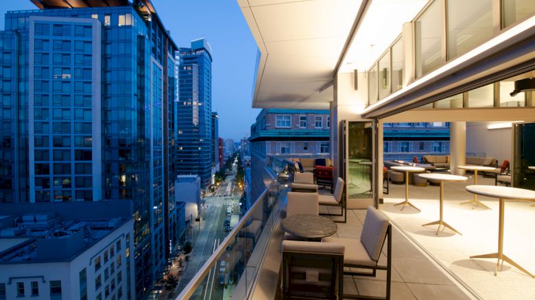A modern rooftop terrace at dusk, with city buildings, elegant furnishings, and a glass railing overlooking the street below.