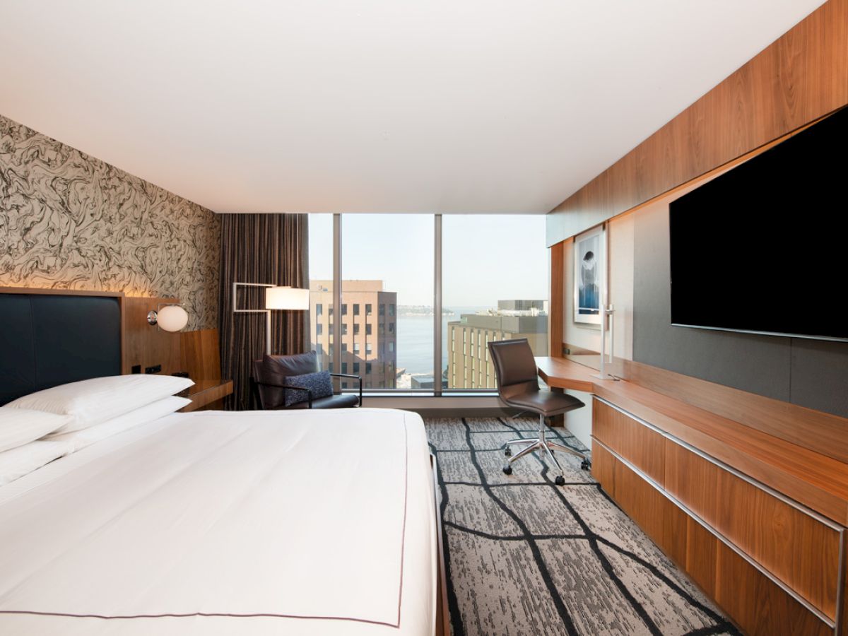 A modern hotel room with a neatly made bed, large TV, desk, and chair. A window reveals a cityscape view and artistic wall decor.