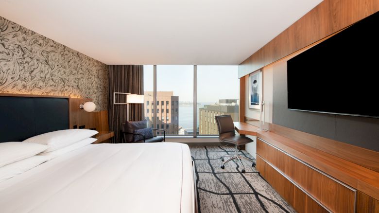 A modern hotel room with a neatly made bed, large TV, desk, and chair. A window reveals a cityscape view and artistic wall decor.