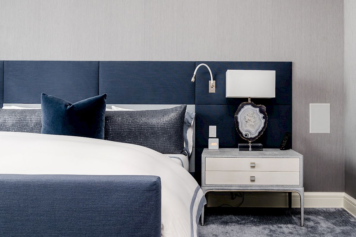 A modern bedroom with a blue and white color scheme, featuring a bed, pillows, a nightstand with a lamp, and a carpeted floor.