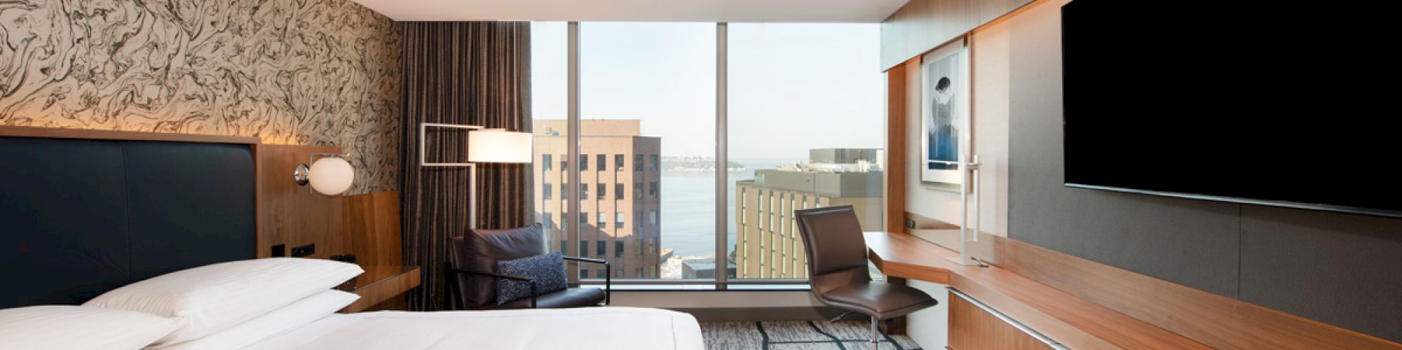 A modern hotel room with a bed, wall-mounted TV, desk, chair, and large windows overlooking the city.